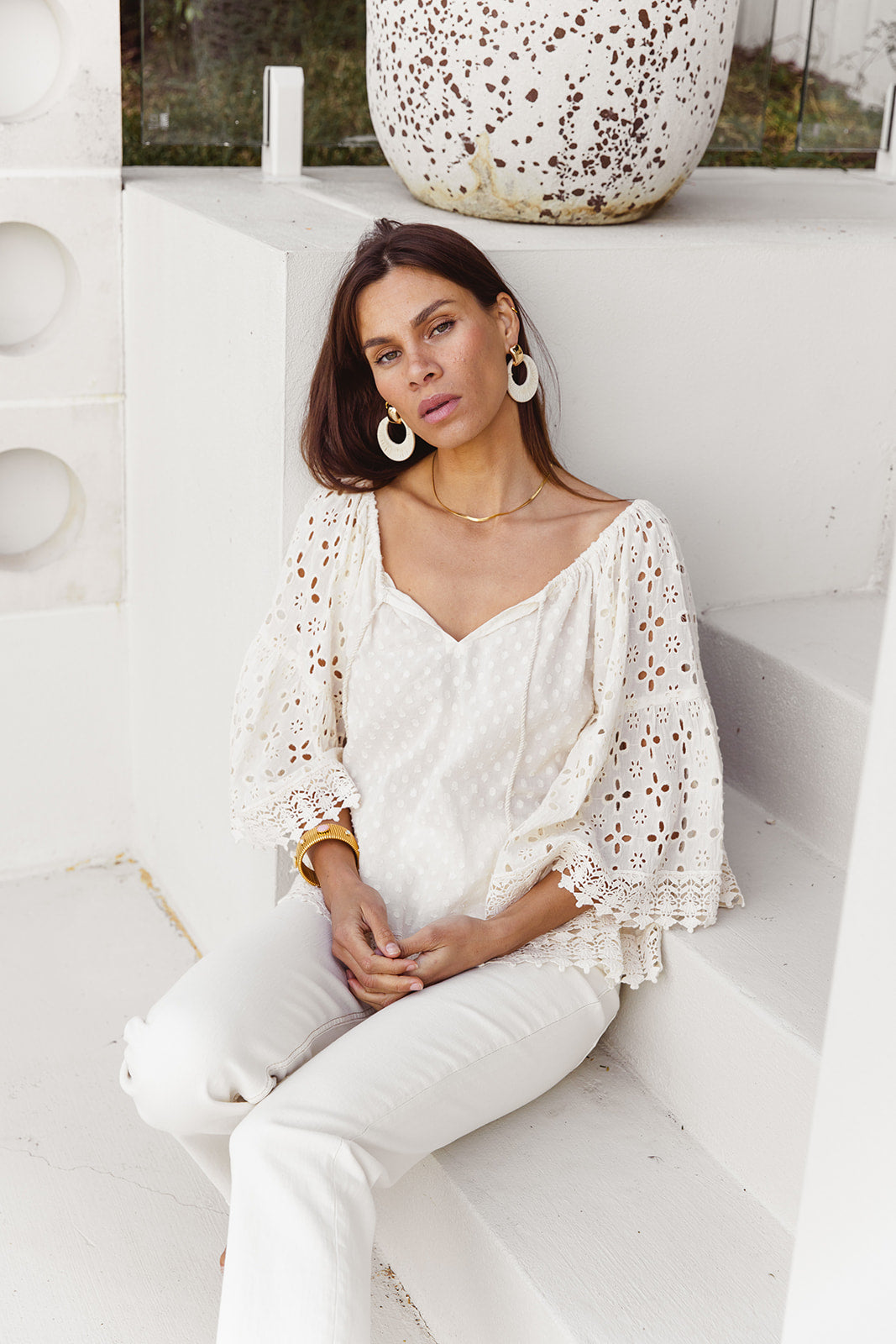 Woman wearing a white lace top and pants sitting on a white bench.
