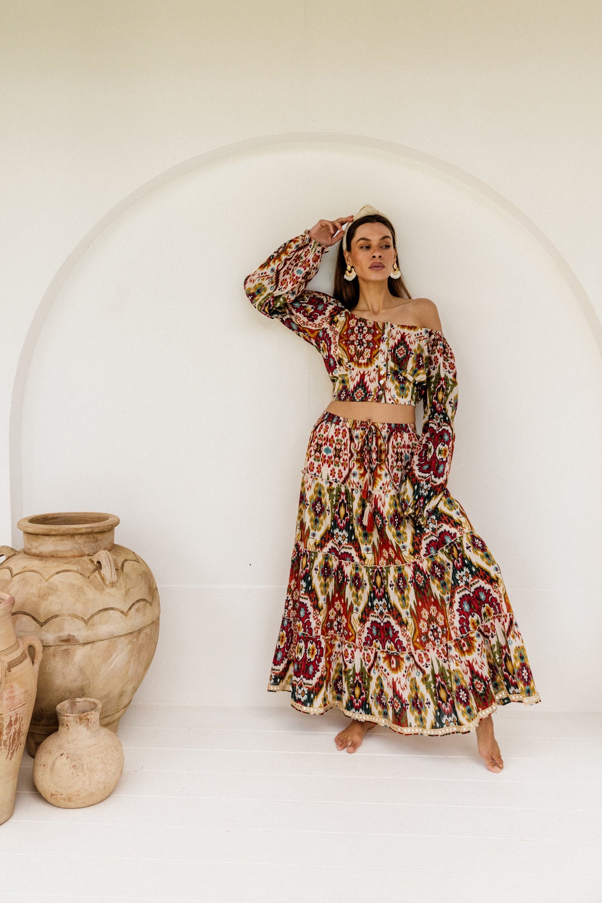 Woman wearing a colorful patterned outfit standing in front of a white archway with ceramic vases.