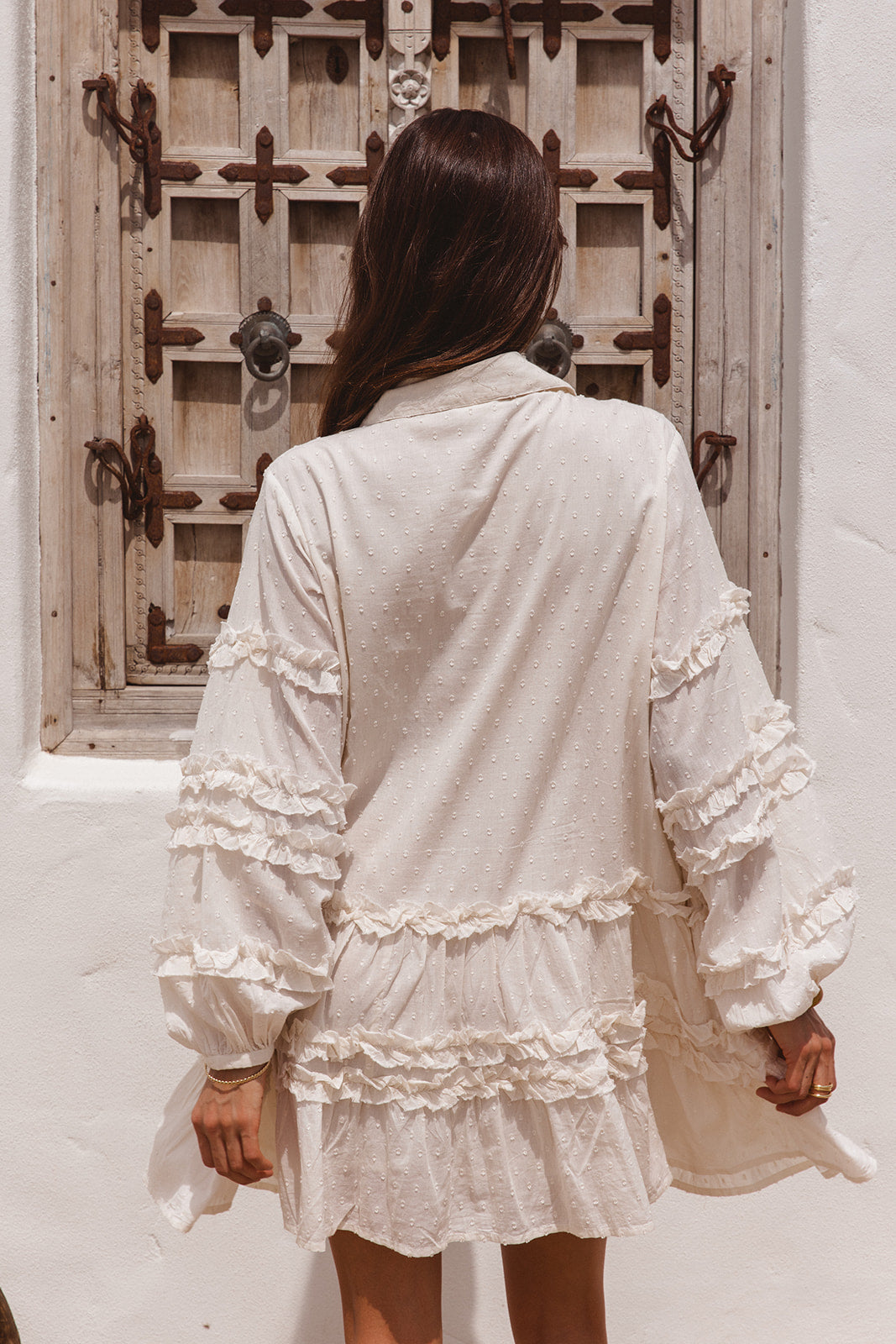 Woman wearing a white dress with ruffles  standing in front of a wooden door.