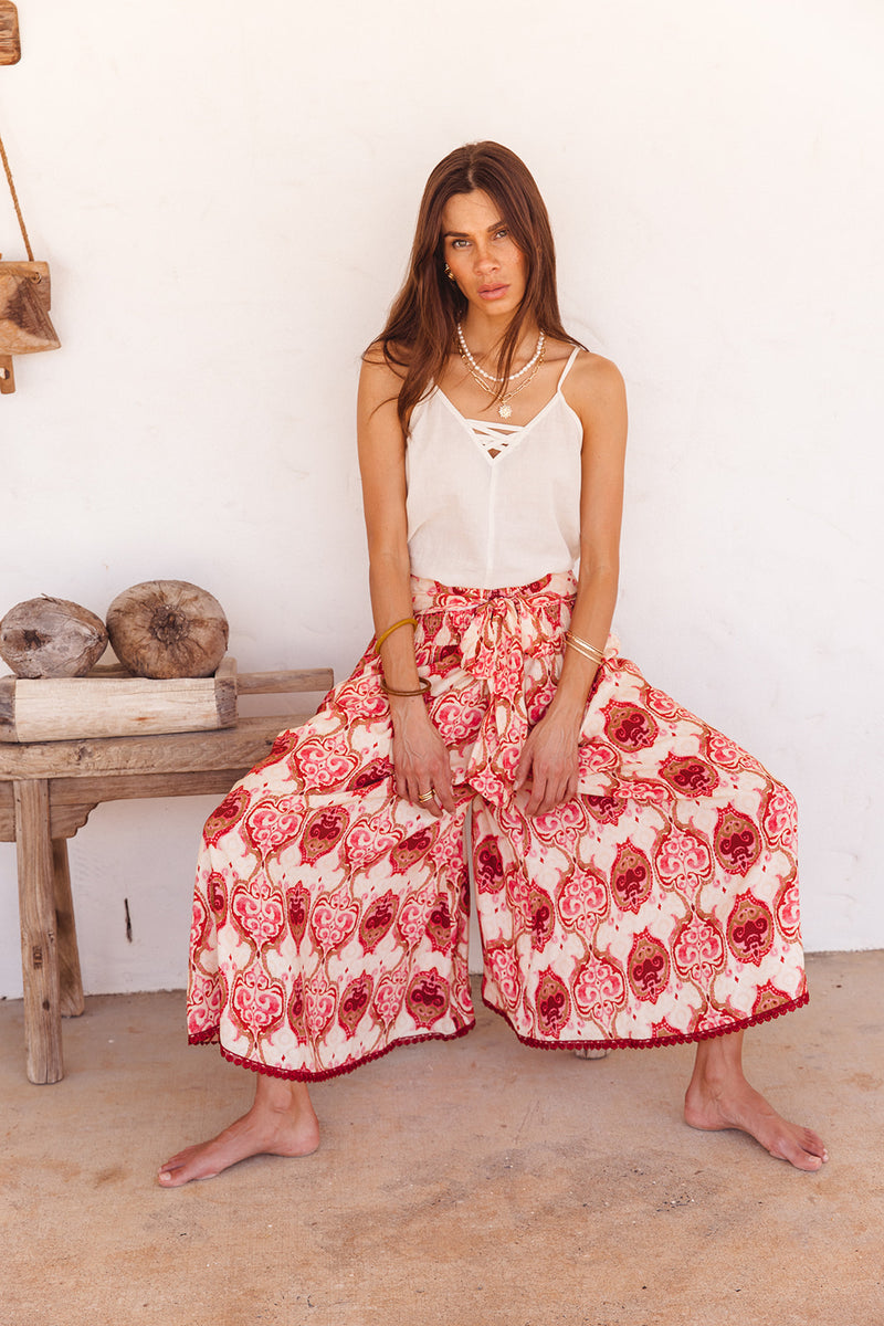 Woman wearing a white top and red floral wide pants sitting on a wooden bench.