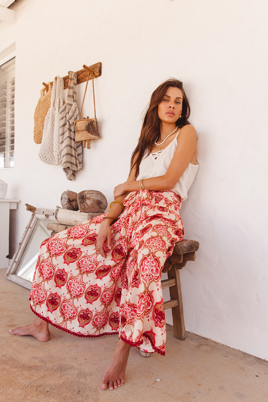 Woman in a white top and red floral wide pants sitting on a wooden stool against a white wall.