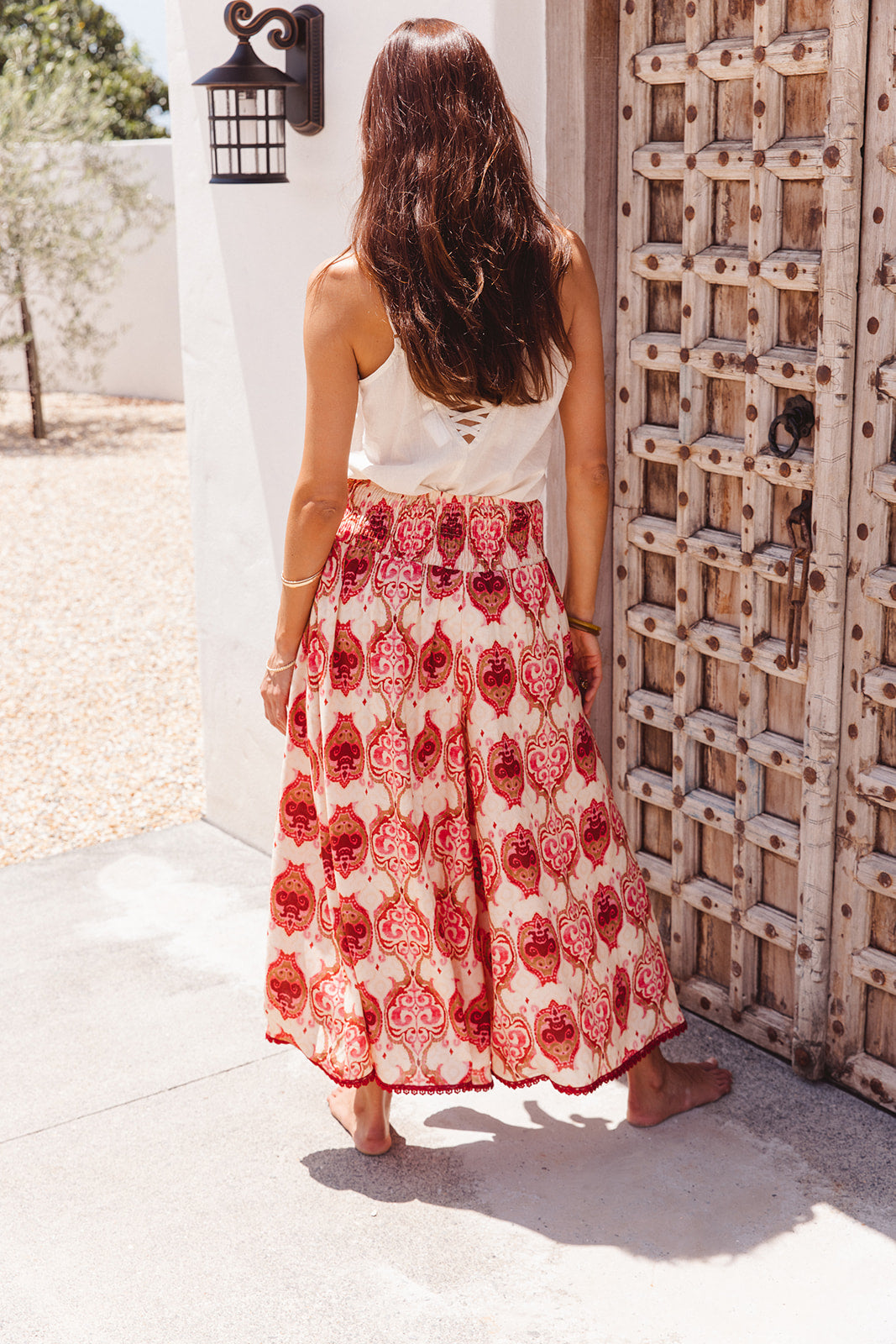 Woman wearing a white top and red patterned wide pants standing in front of a wooden door.