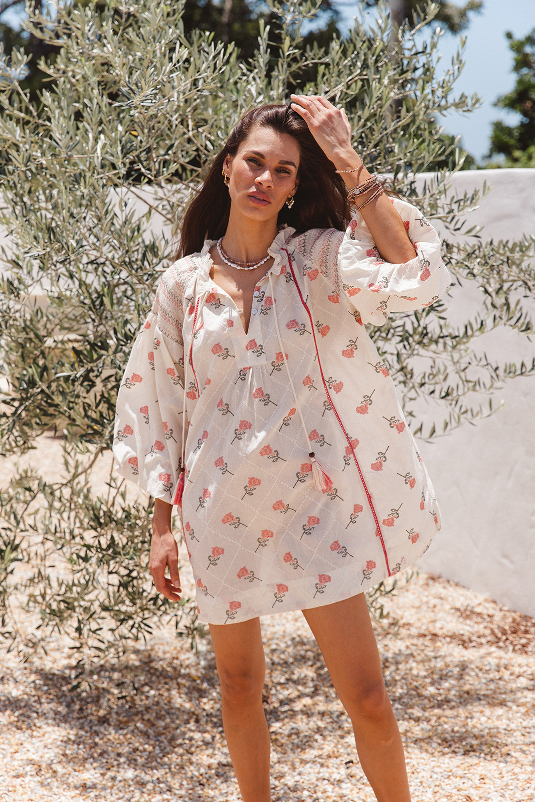 Woman in a floral dress standing outdoors with trees and a white wall in the background