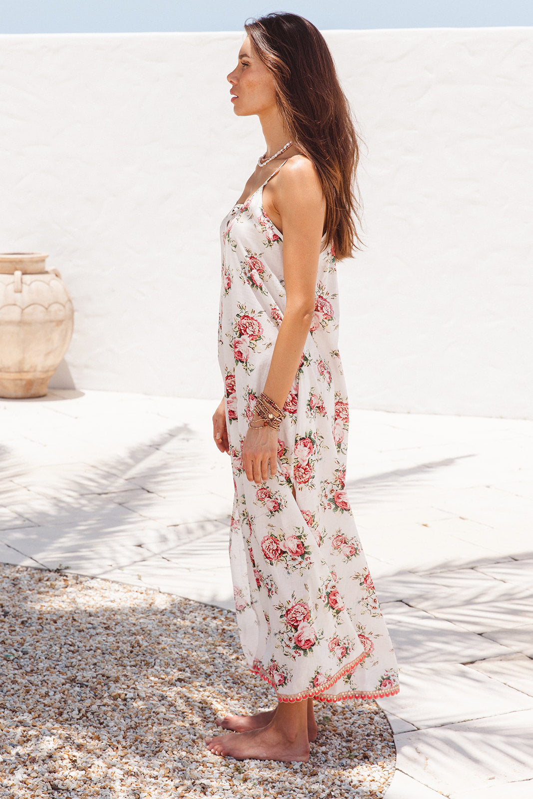 Woman in a floral dress standing on a pebbled beach with a white wall and vase in the background.