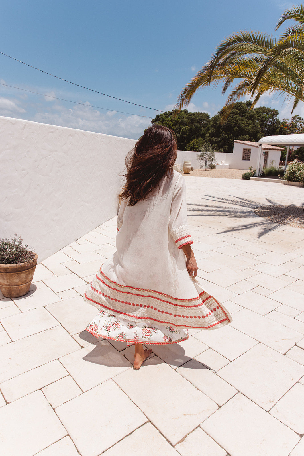 Woman in a white robe with red patterns walking on a tiled patio with palm trees in the background.