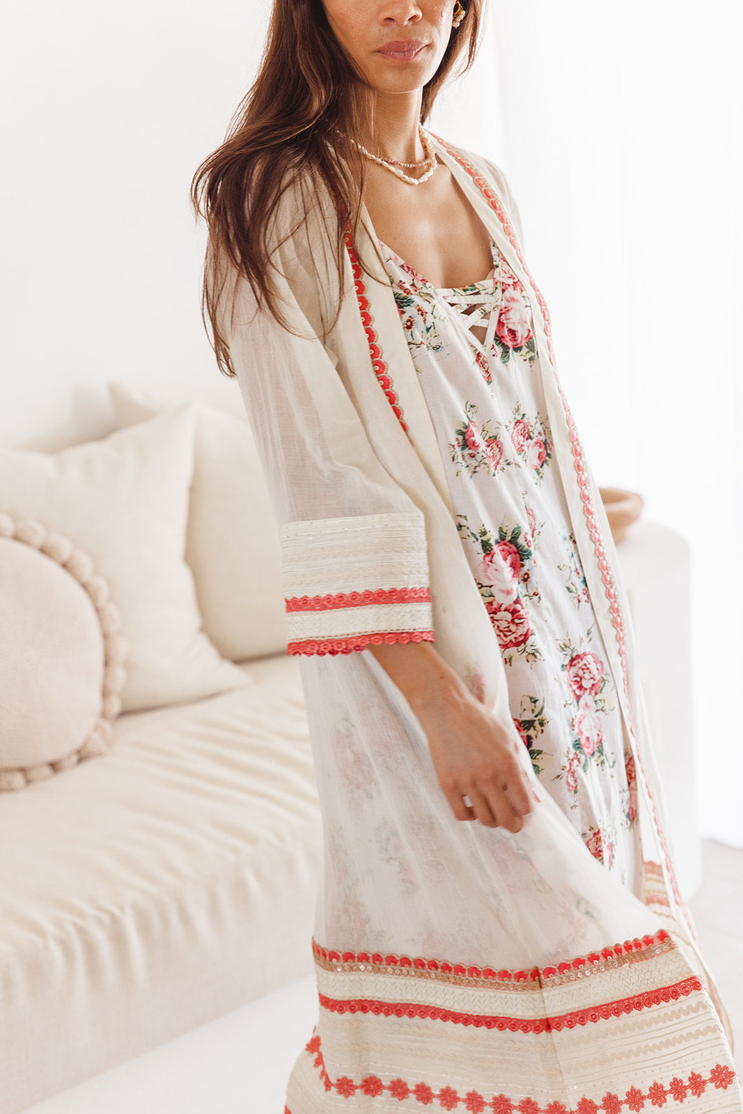 Woman wearing a floral dress with red accents standing against a white couch.