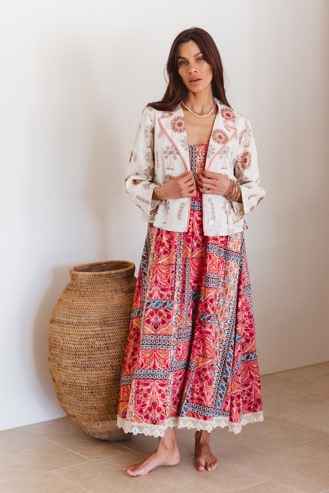 Woman wearing a white jacket and red patterned skirt standing against a plain wall with a woven basket.