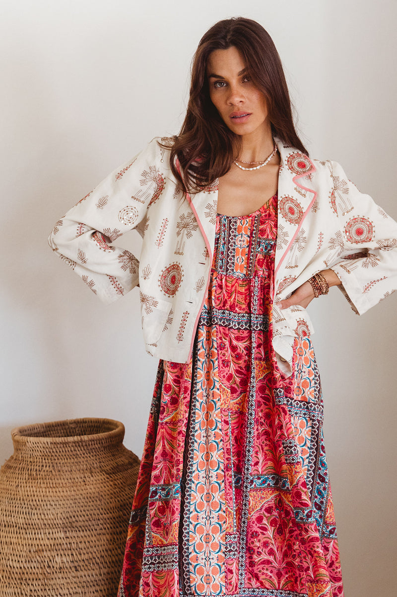 Woman wearing a colorful dress with a white embroidered jacket, standing against a plain background.