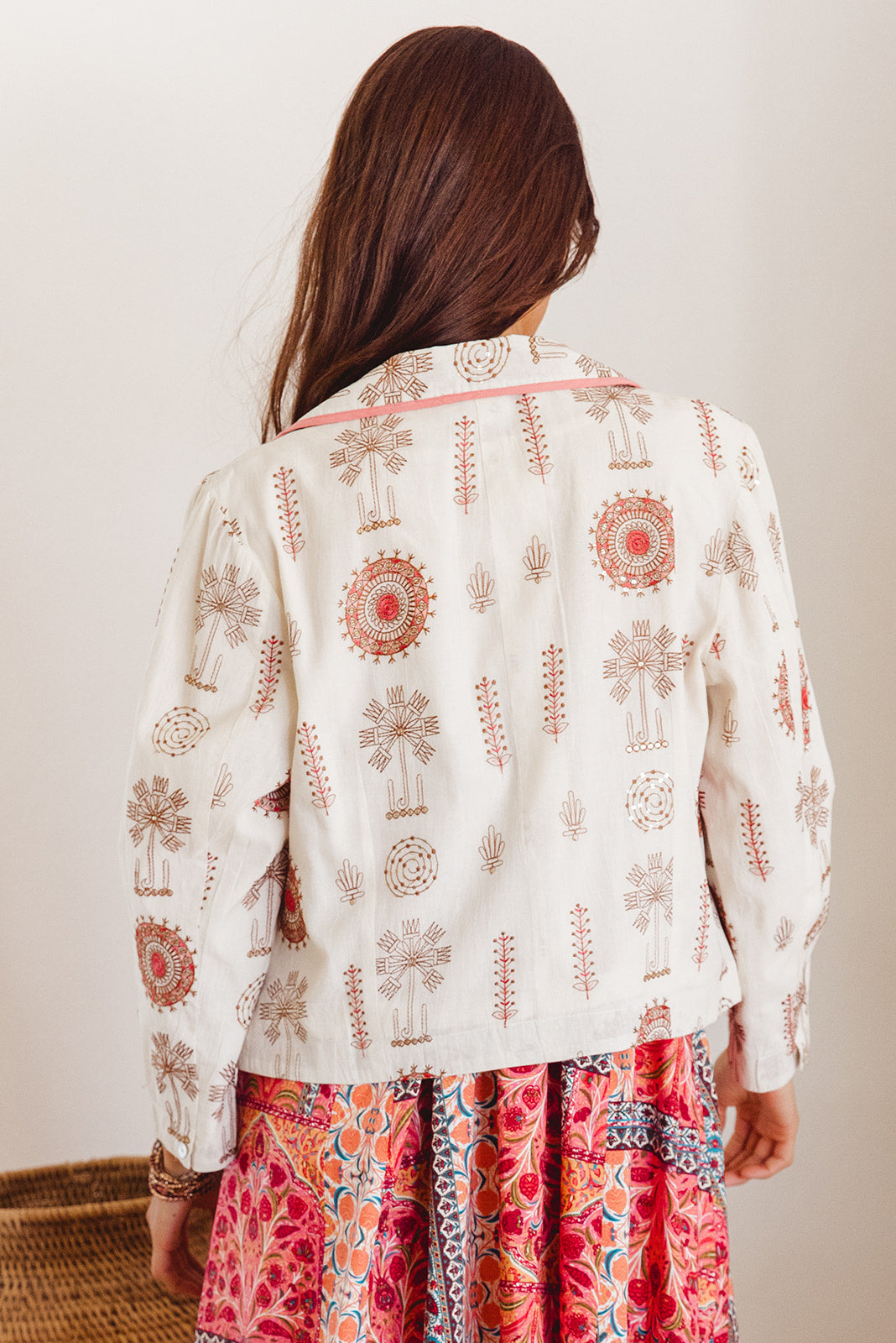 Person wearing a white jacket with red and gold patterns and colorful dress against a plain background