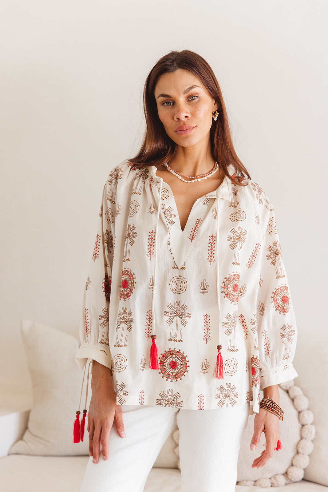 Woman wearing a white blouse with red patterns and tassels in a neutral setting