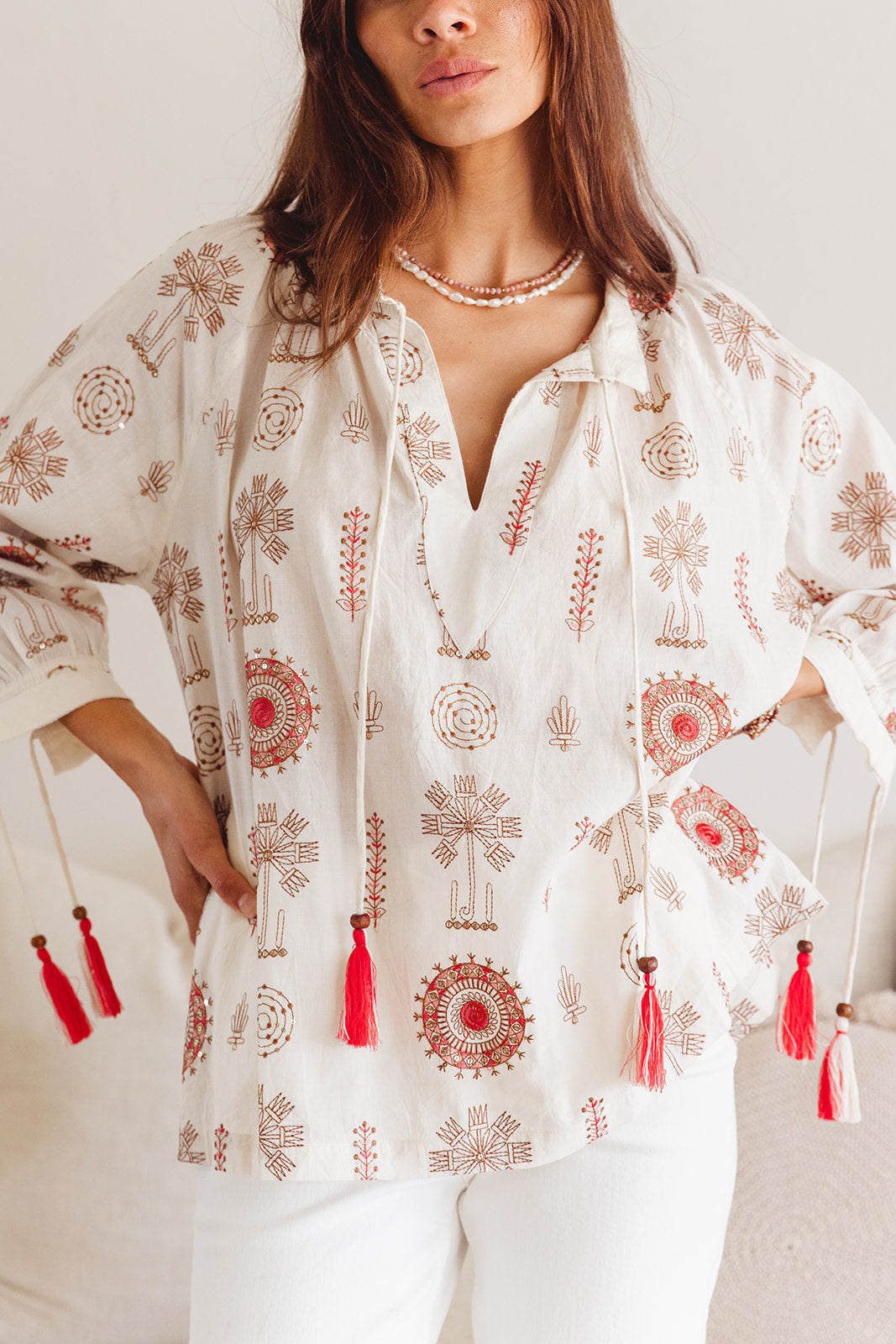 Woman wearing a white blouse with red floral patterns and tassels, standing against a plain background.