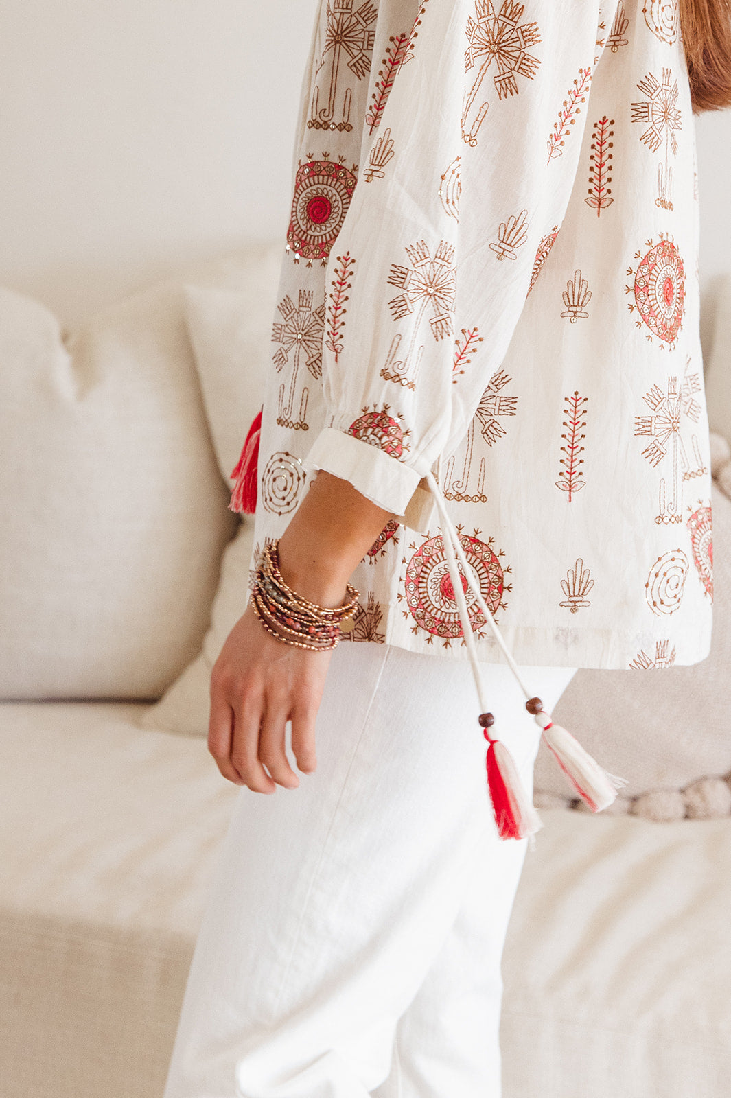 Person wearing a white outfit with red patterns and tassels, standing by a beige couch.