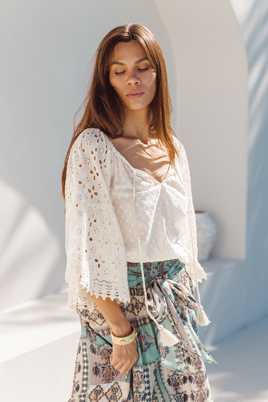 Woman wearing a white lace top and patterned skirt against a white wall.