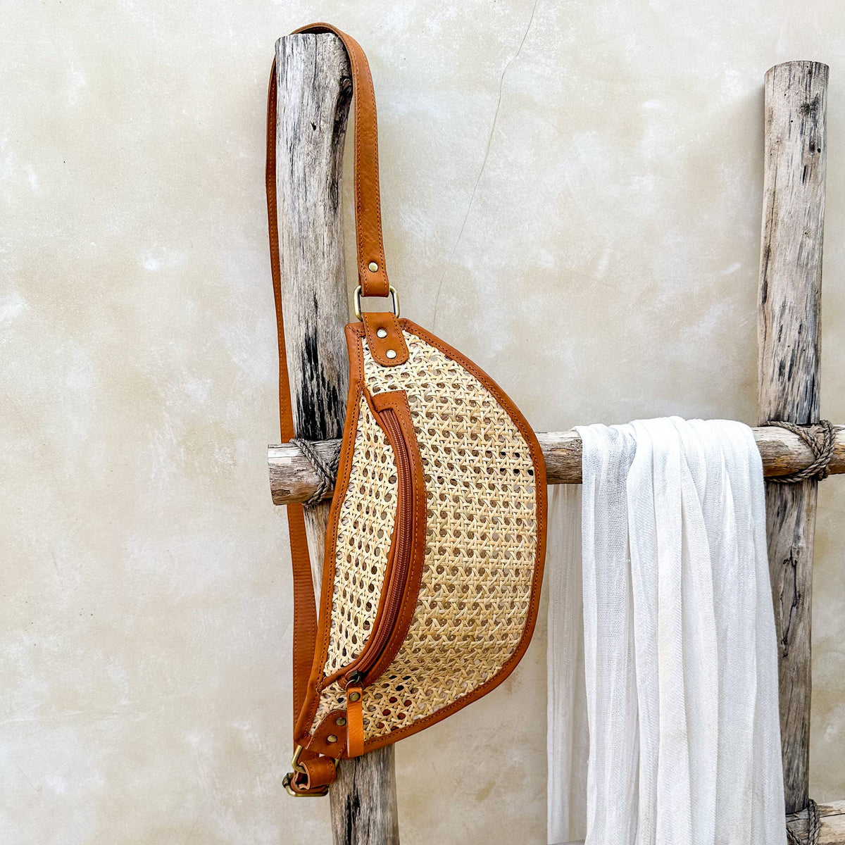 Straw bag with brown leather strap on a wooden rack against a textured wall.